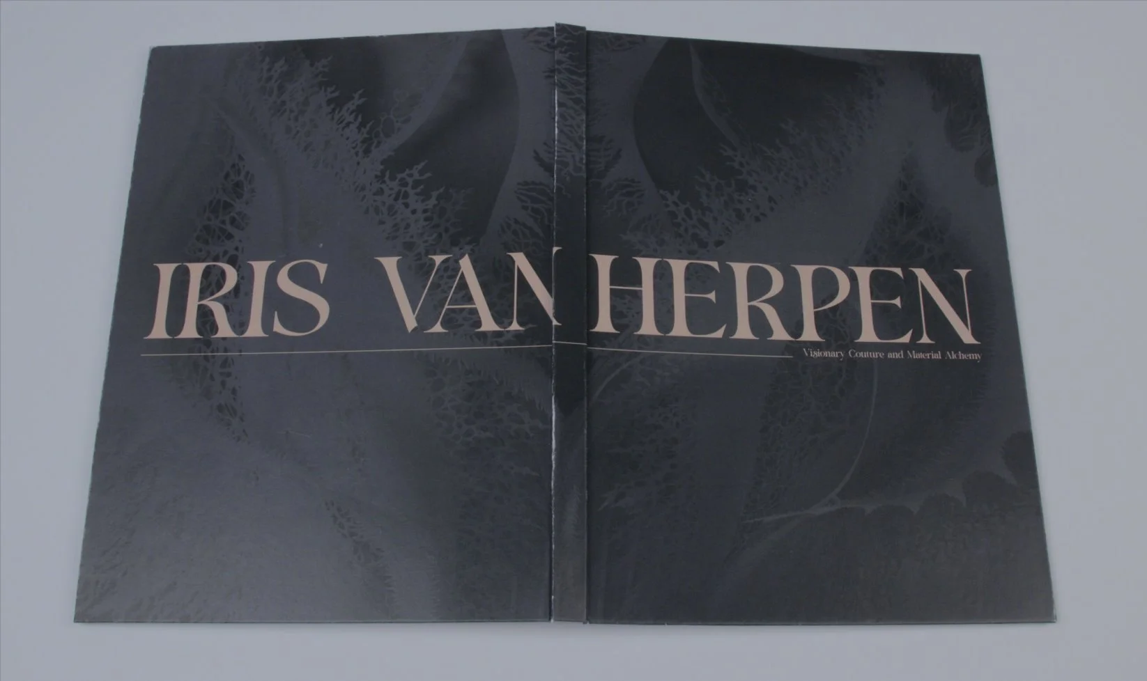 The catalog cover laid open, showing the full IRIS VAN HERPEN title in cream against deep black with darkened couture imagery in the background